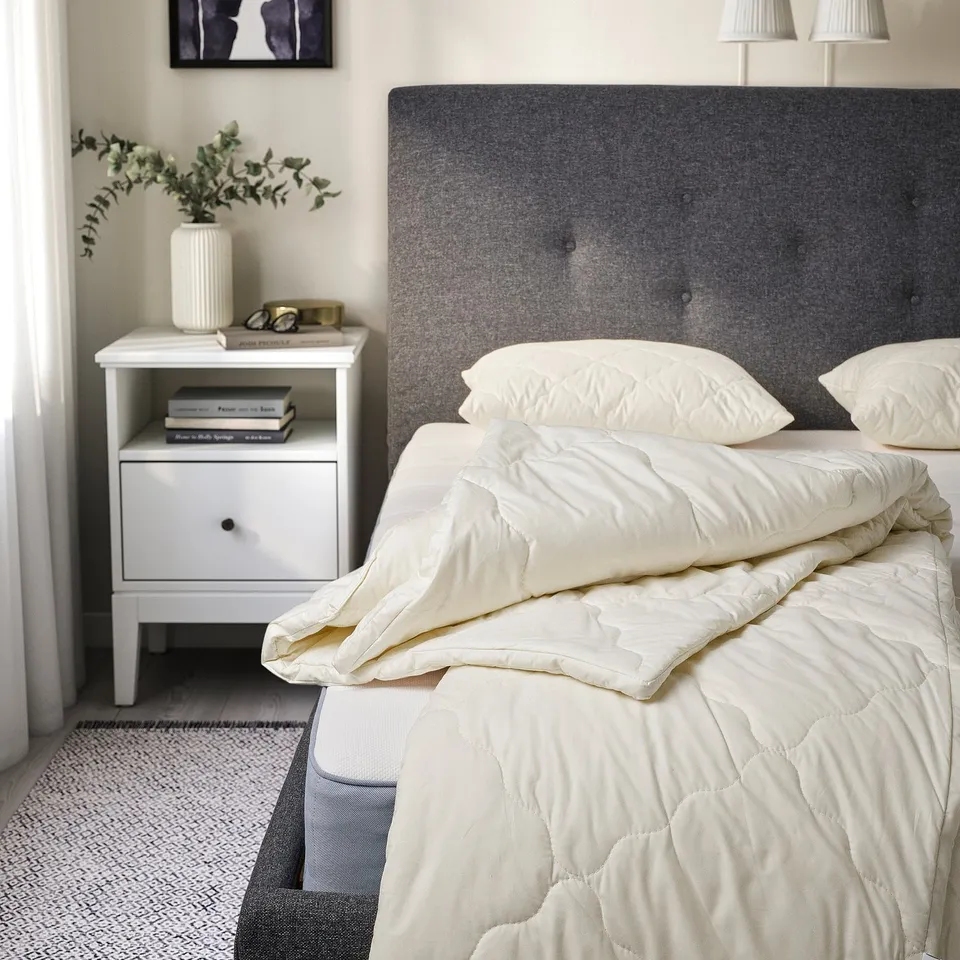 Minimalist bedroom with grey duvet, two white pillows, grey headboard, bedside table, and floral vase.