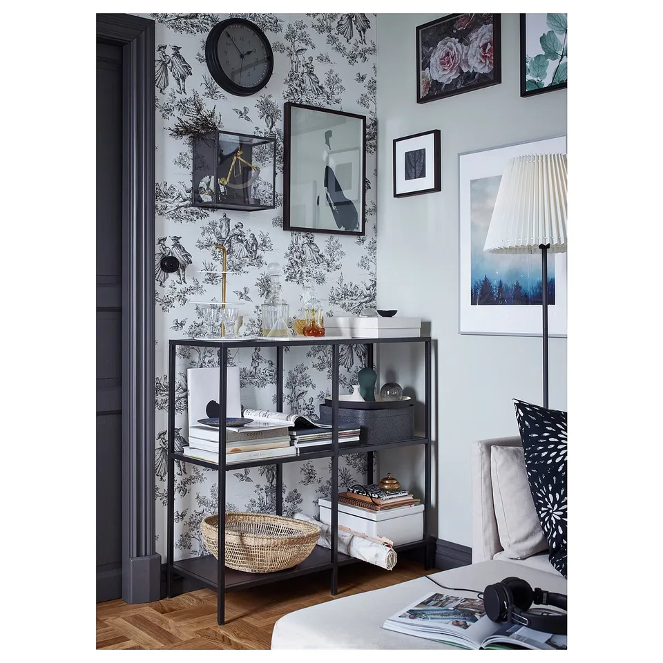 Black-brown glass bookshelf in bedroom with artwork, clock, and floral wallpaper.