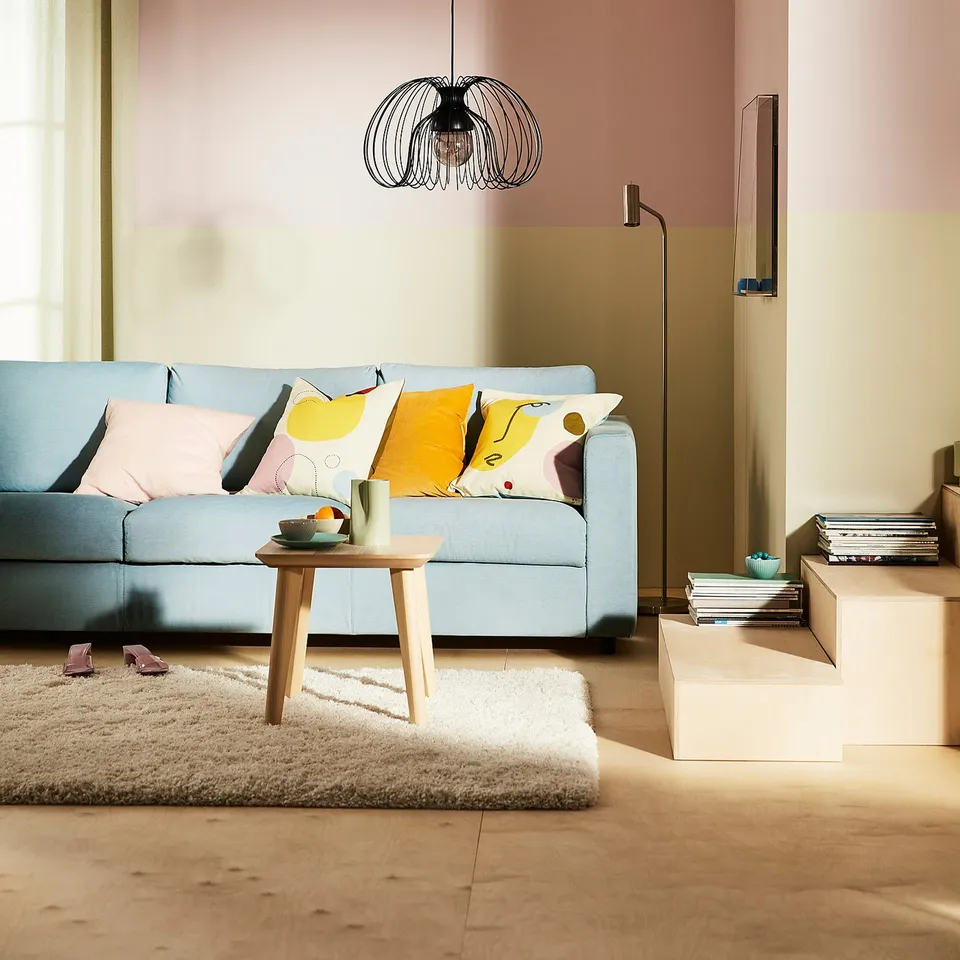 A modern living room with a light blue VIMLE sofa, colourful pillows, wooden coffee table, fluffy white rug, and a black pendant light.