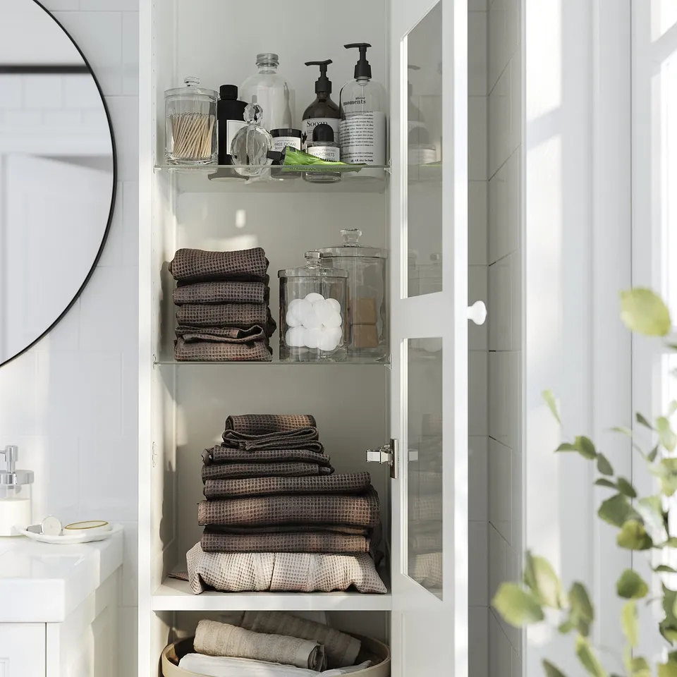 White wall-mounted bathroom cabinet with glass door, showing towels and bottles.