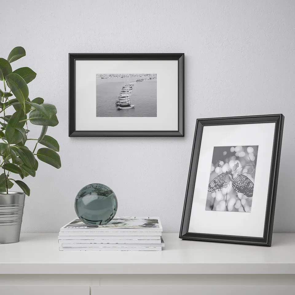 Modern decor setup with KNOPPÄNG picture frames showing sailors and butterflies, a reflective sphere, and a green plant on a white surface.