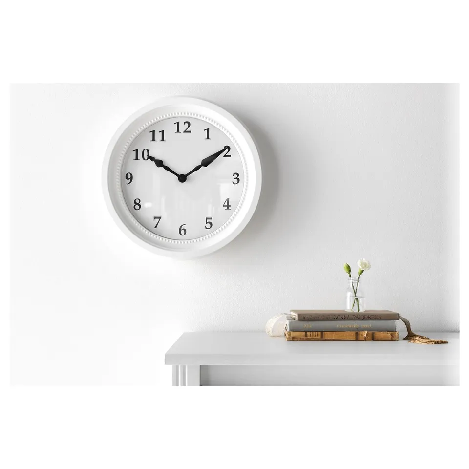White round wall clock with black numbers and hands, silence quartz movement, flanked by a small vase with white flowers on a white shelf.