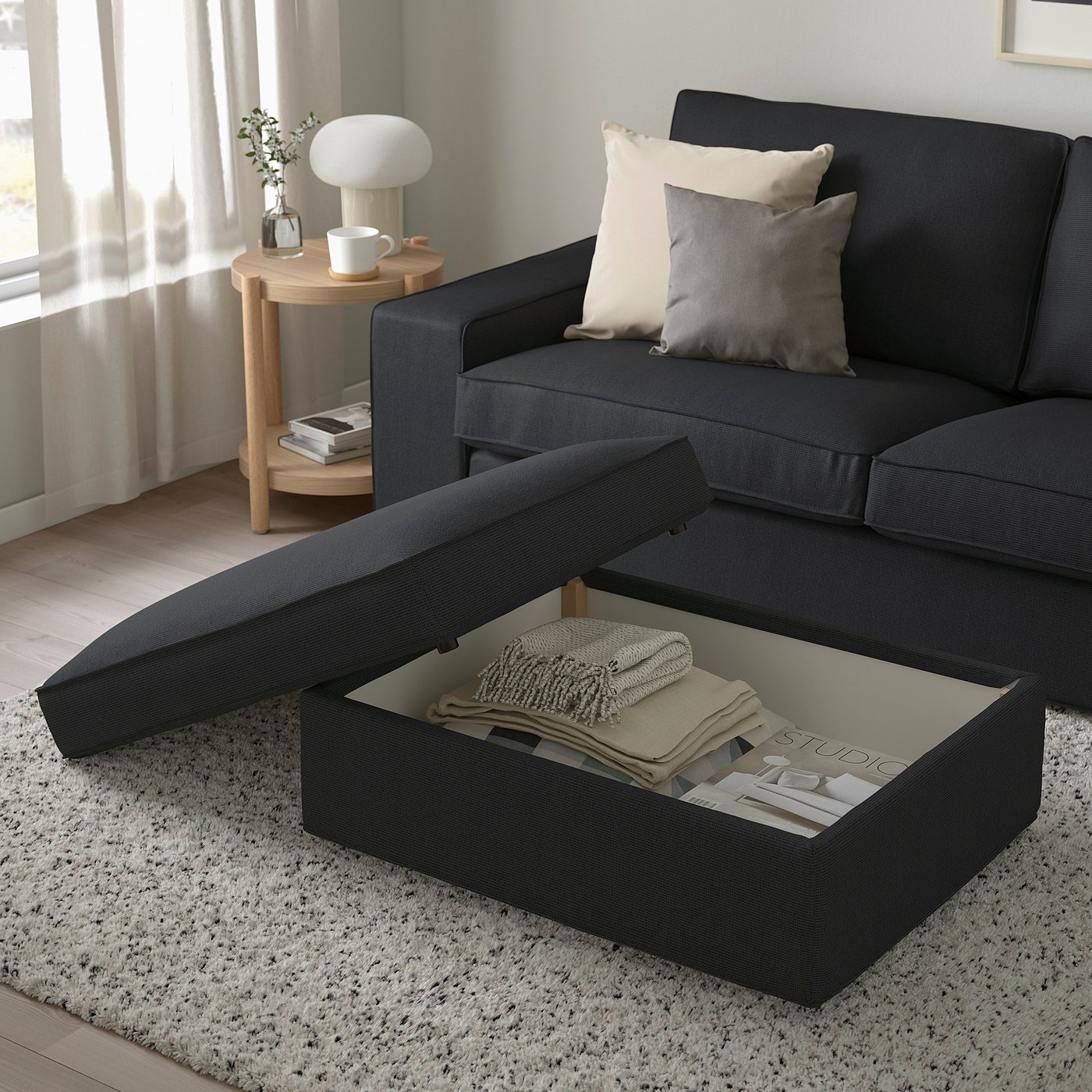 KIVIK footstool with blanket & magazines, beside modern grey couch on patterned rug, near table with lamp & vase.