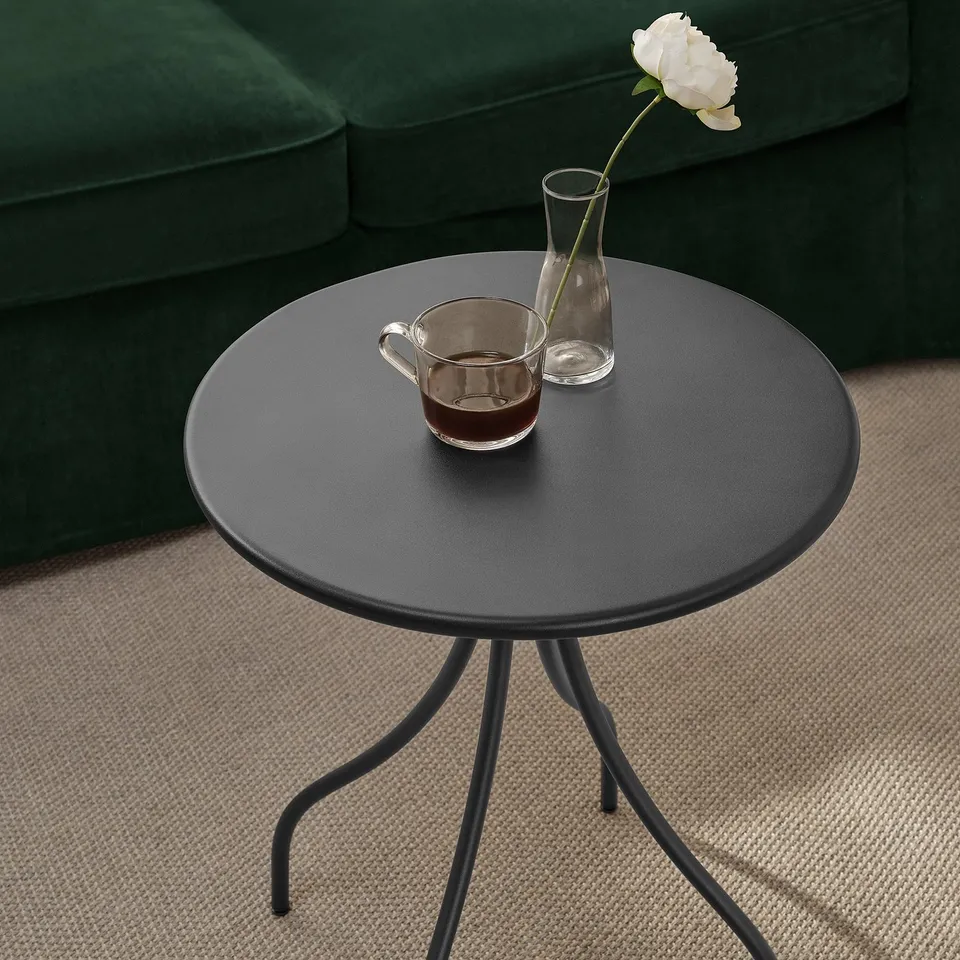 Small round black side table with thin legs holds a cup of coffee and a small vase with a single white flower. Perfect for a cosy corner.