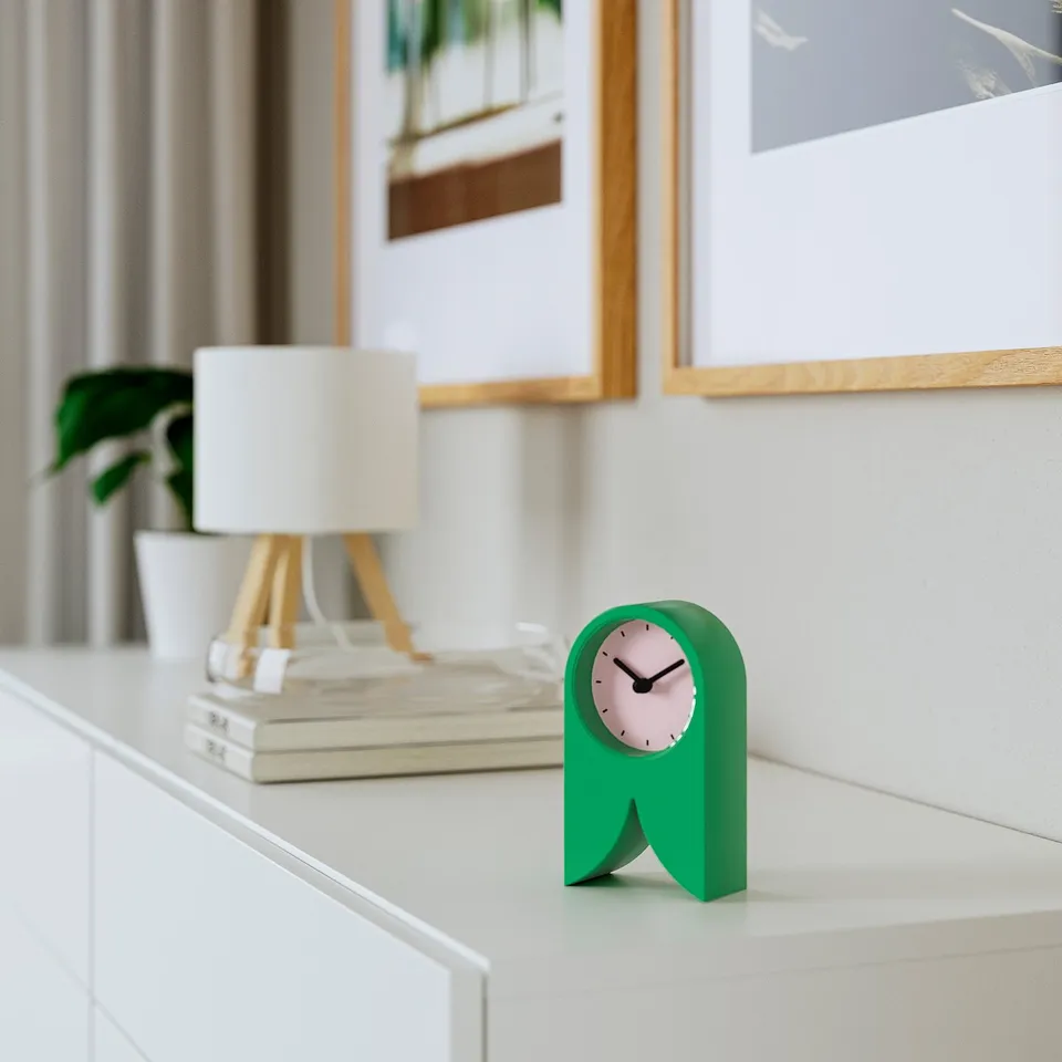 Green FLODKRÄFTA clock on white table, minimalist design, silent quartz mechanism, with books and lamp.