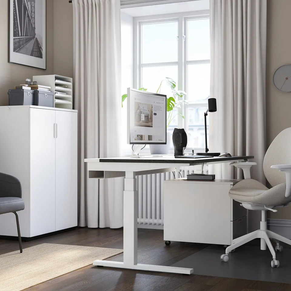 Modern home office setup. White sit/stand desk centre, large screen, chair. Cabinet, art, plant, rug. Window with sheer curtains.
