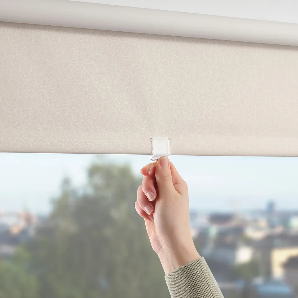 Hand lifting beige roller blind, slightly open with city view; smooth, light-blocking function.