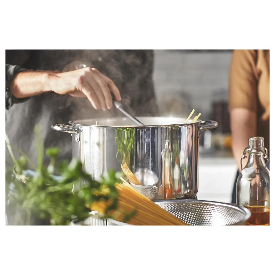 Cooking scene with stainless steel pots, pastas, green herbs, and person stirring.