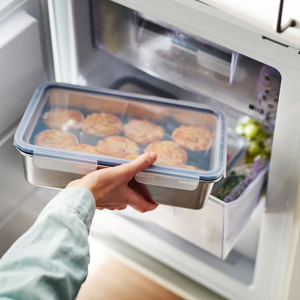 Hand holding stainless steel container with food inside open fridge.