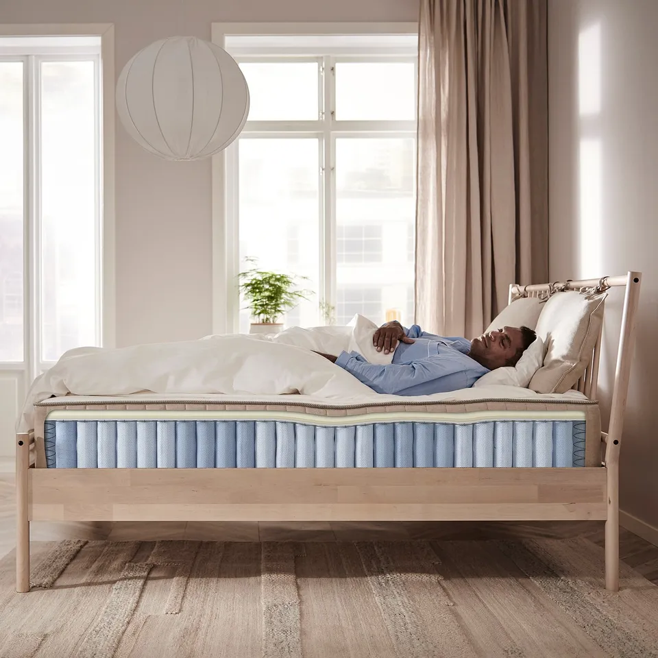 Person resting on a white, blue, and brown VATNESTRÖM mattress on a wooden bed next to a window with curtains.