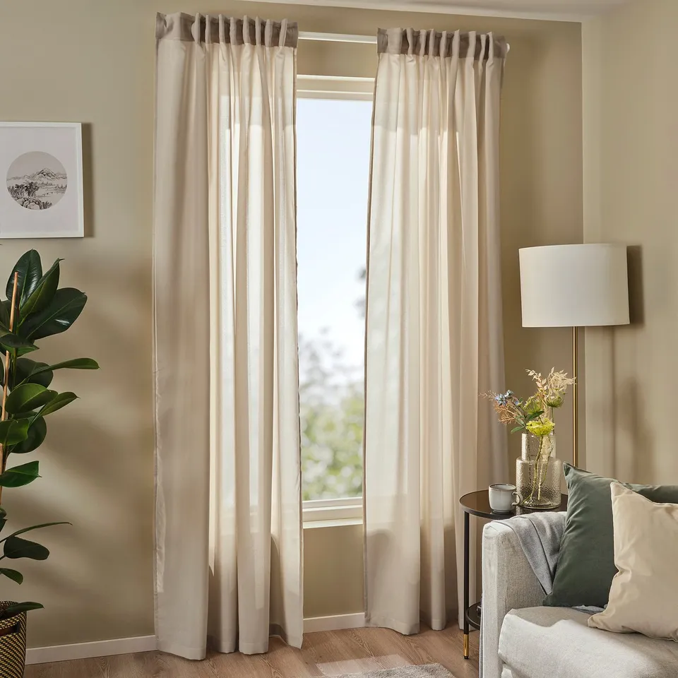 Modern living room with large beige ROSENROBINIA sheer curtains framing a wide window, complemented by a cosy corner sofa and tall indoor plant.