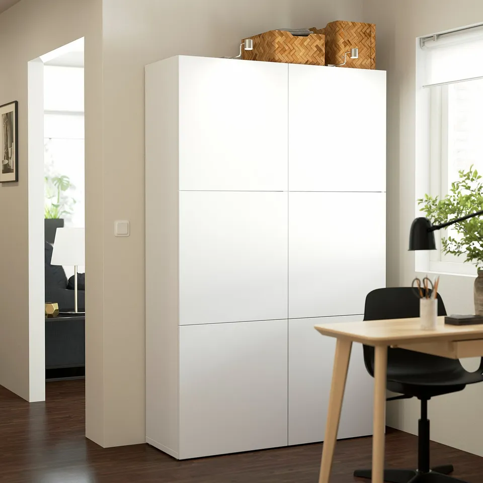 Large white BESTÅ cabinet stands tall near a window, taller than the desk beside it, with wicker baskets on top and sleek, minimalist design.