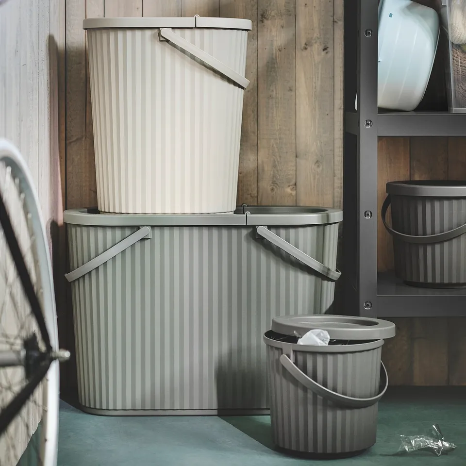 Three grey DAMMÄNG bins - large, medium, small - against wooden wall and metal storage on teal floor. Designed for small item waste sorting.