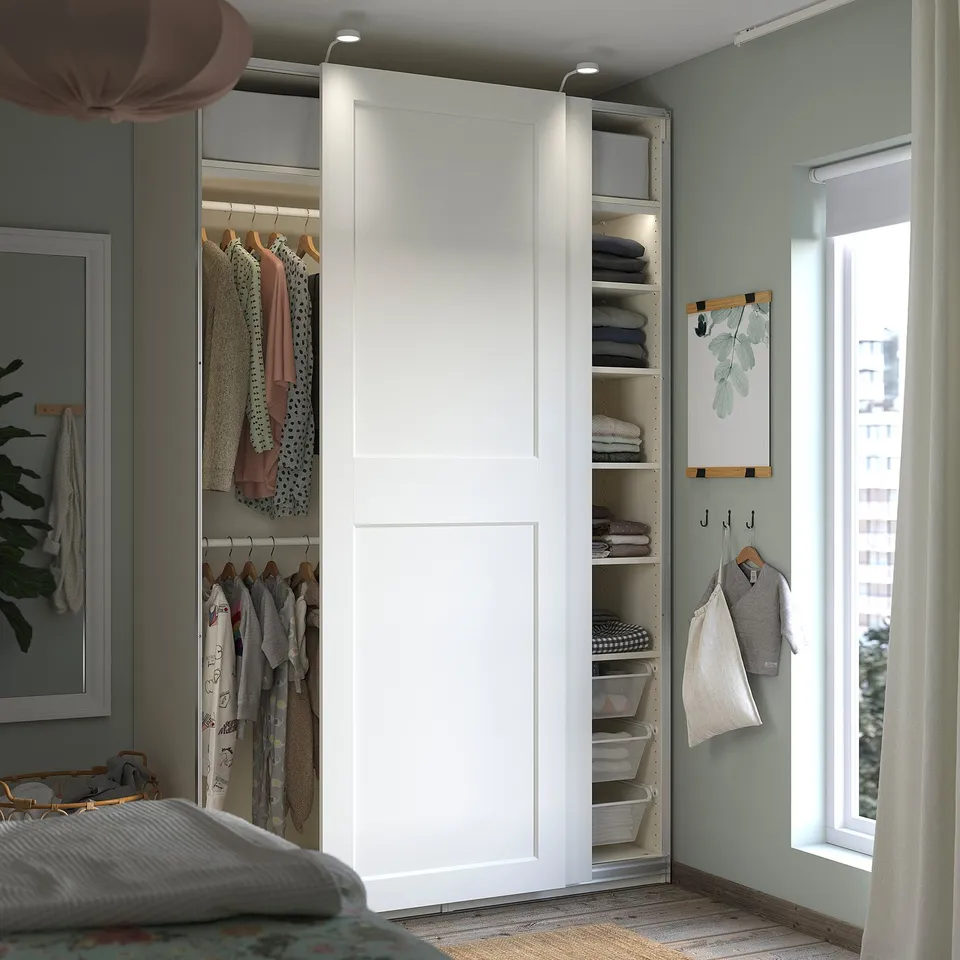 A white IKEA PAX wardrobe with sliding doors, containing shelves, hangers, and mesh baskets, organised with clothes.
