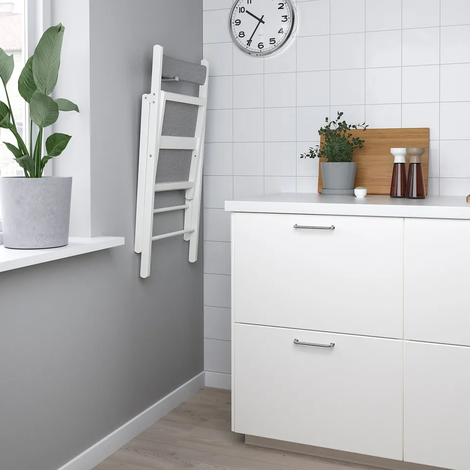 Modern kitchen with white cabinet, foldable chair, potted plant, clock, and utensils.