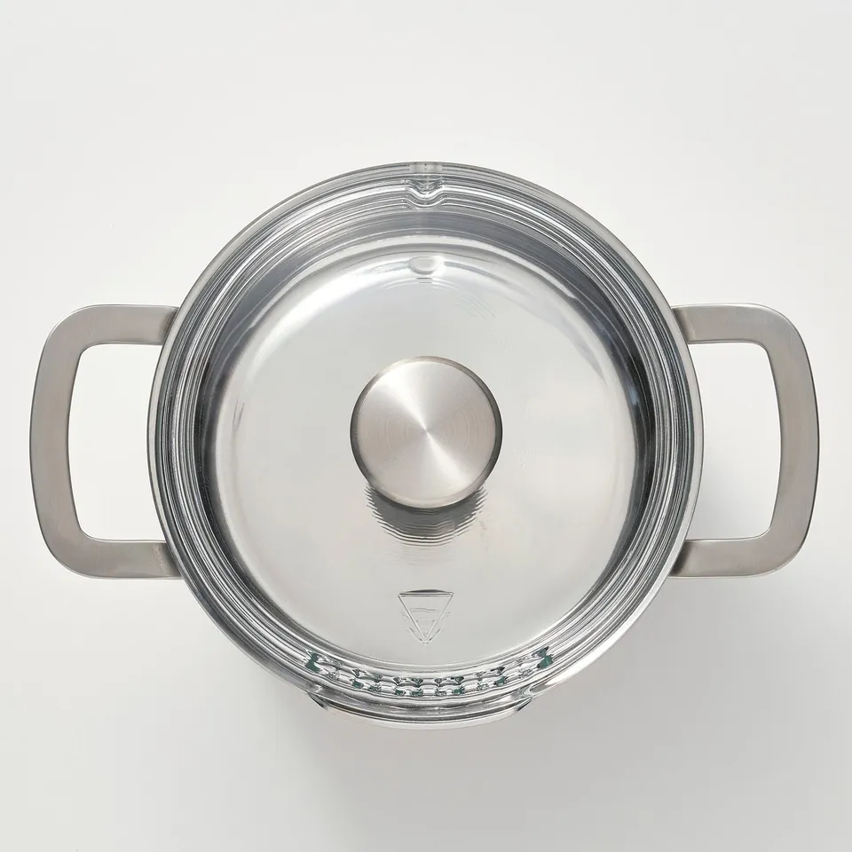 Silver saucepan with clear glass lid and metal handles viewed from above, reflecting light, demonstrating high-quality stainless steel construction.