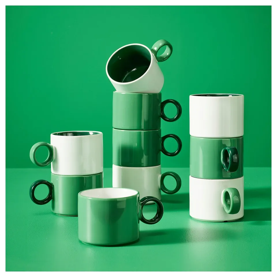 Stacked green and white PIGGÅL mugs on a green background, featuring contrasting colours and a simple design in stoneware.