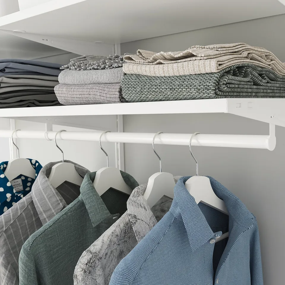 A closet with white shelves and hanging rods. The top shelf holds folded blankets. Below, clothes hang on white hangers.