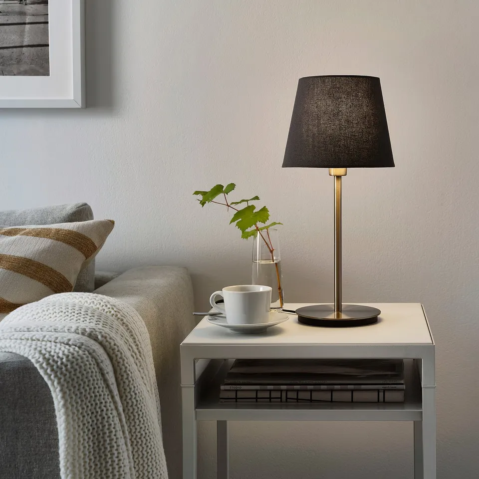 Small SKAFTET black lamp on side table, beside cosy grey couch with white blanket. Books and plant nearby.