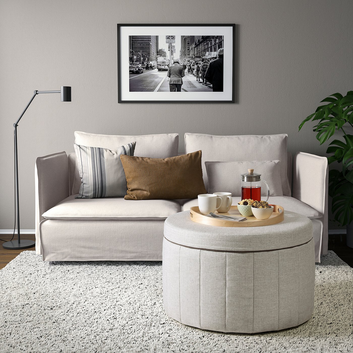 Modern living room with light grey l-shaped sofa, round ottoman, plants, and black & white city photo.