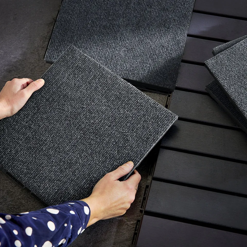 Assembling RUNNEN black floor tiles from recycled materials on wooden slats; wears blue polka-dot shirt.