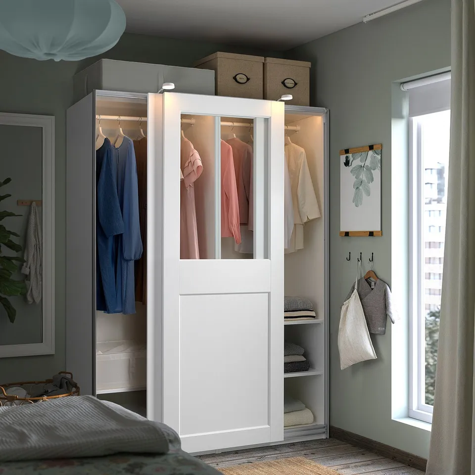 White PAX wardrobe with glass doors, clothes inside, next to window and bed. Mirrored and lit.