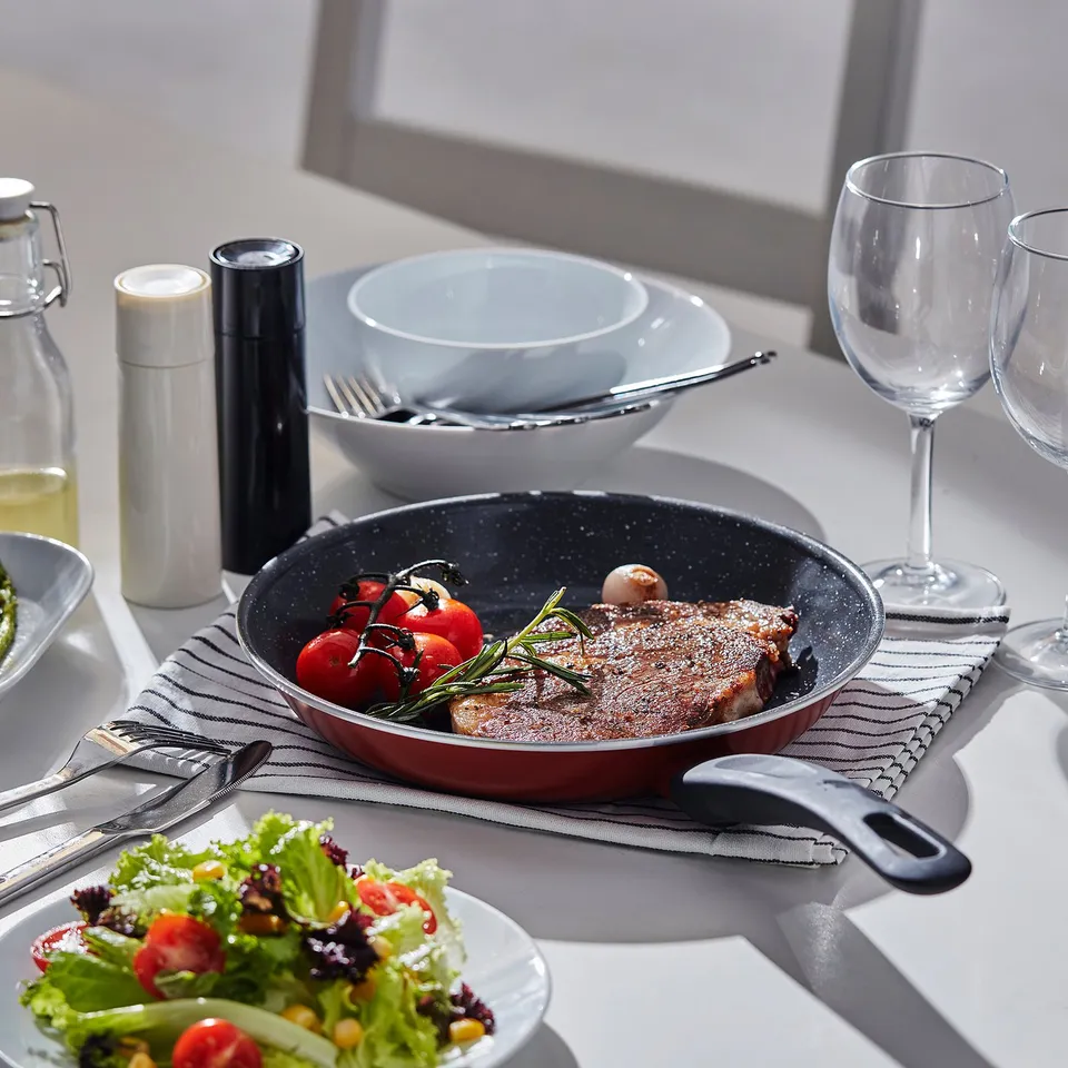 Table set with frying pan, plates, glasses, salad, and utensils. A fried meat dish with tomatoes and herbs is in the pan.