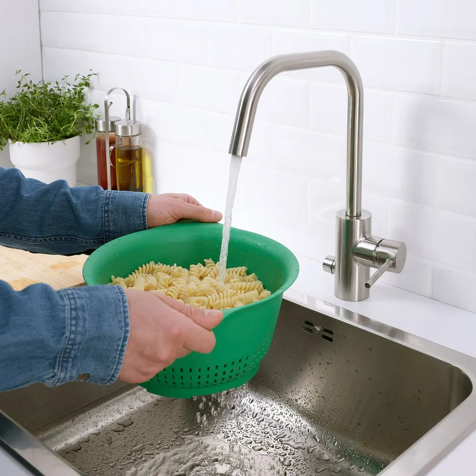 Person rinsing cooked pasta in green colander under modern kitchen tap.