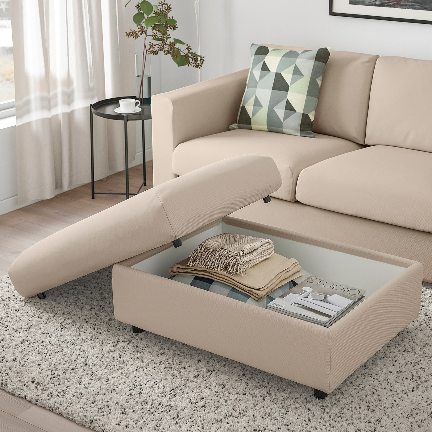 Living room with beige VIMLE sofa, footstool with open storage showing blankets, magazines, and wheels. Side table with cup and plant.
