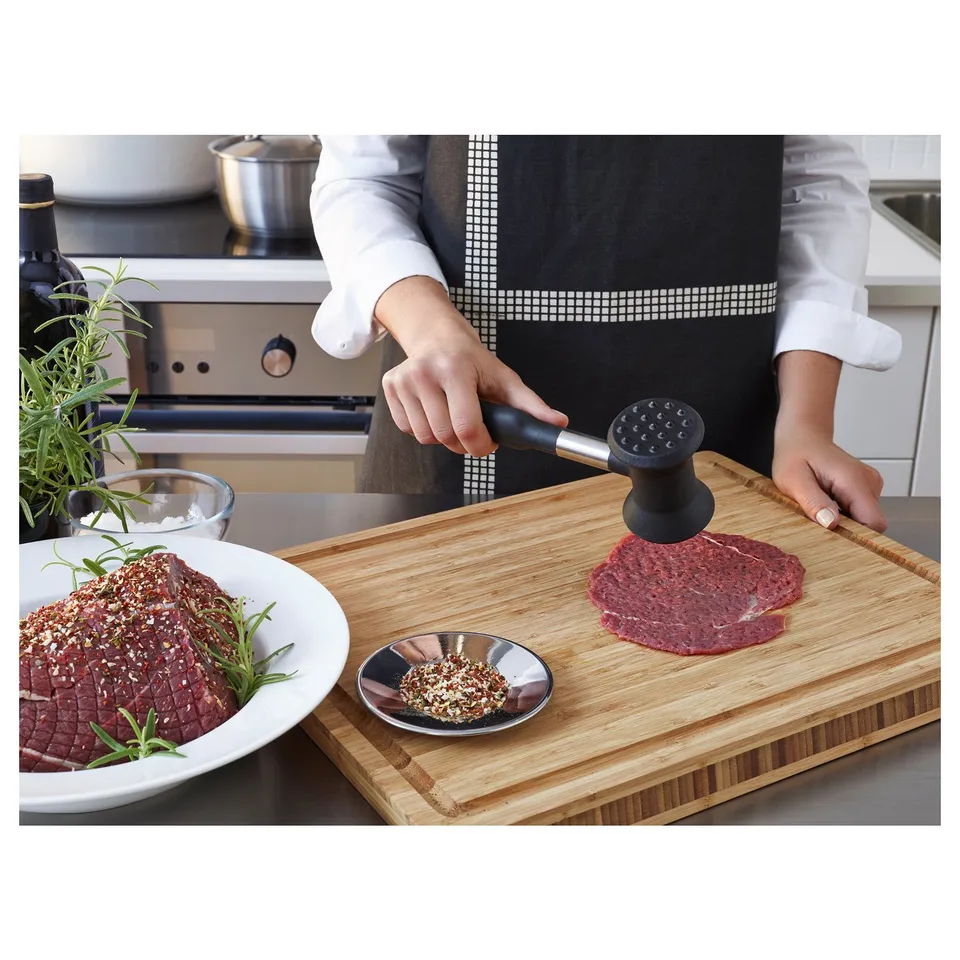 Chef using a black-handled meat mallet on a wooden cutting board with a seasoned meat patty and bowl of spices nearby.