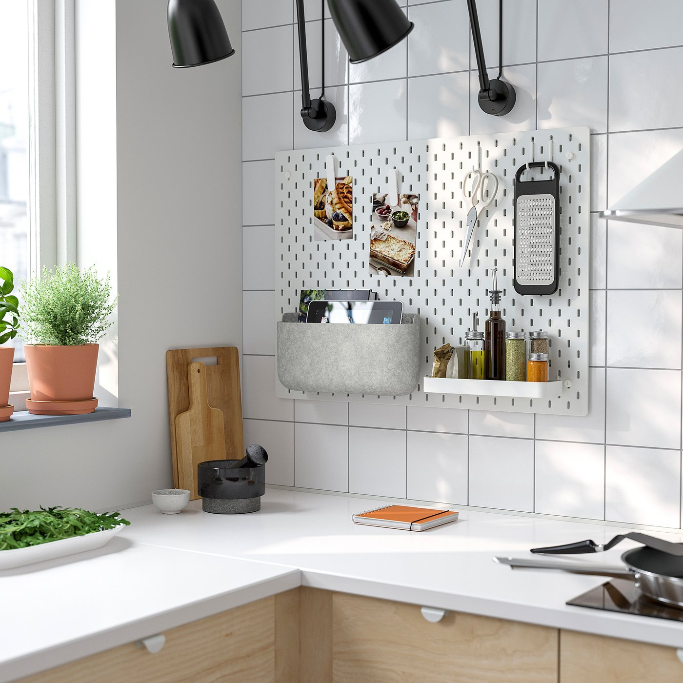 Modern white kitchen with SKÅDIS pegboard organiser holding utensils and spices near the window sill, accompanied by black lamps and wooden elements.