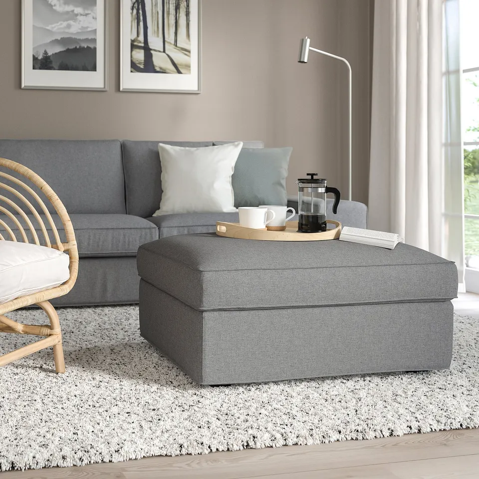 Modern living room with grey KIVIK storage footstool, large for seating or storage, near a blue sofa, a wicker chair, and a white rug.