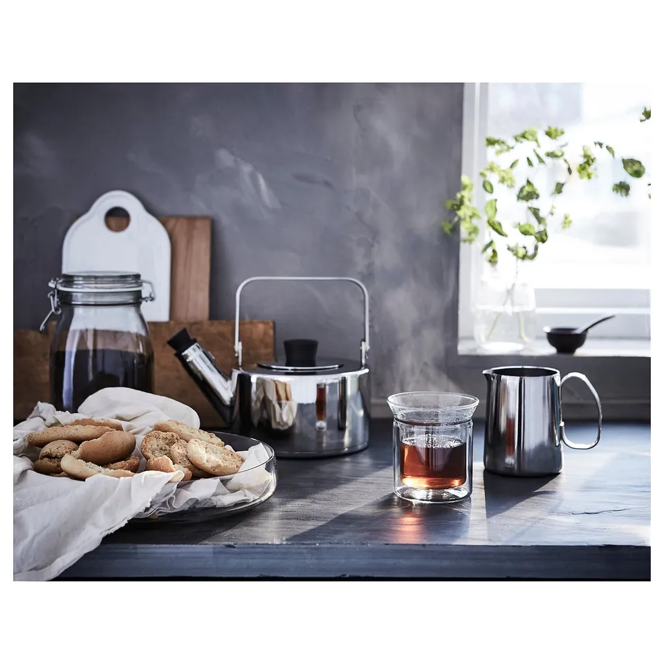 Countertop with MÅTTLIG silver jug, cookies, jar, kettle, and flowers. Sunlight streams through the window.