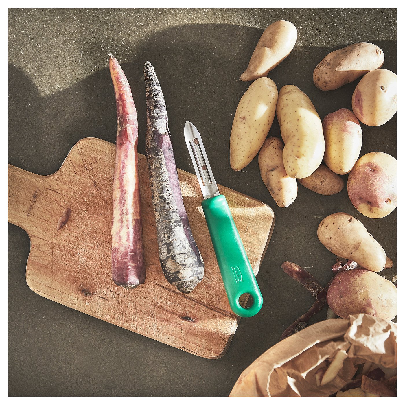 A wooden cutting board holds two root vegetables and a green-handled stainless steel peeler. Several potatoes and a paper bag are also present.