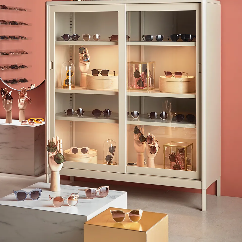 Modern eyewear store with IDÅSEN white metal cabinets displaying stylish sunglasses, pink and gold accessories, and framed pictures on peach walls.