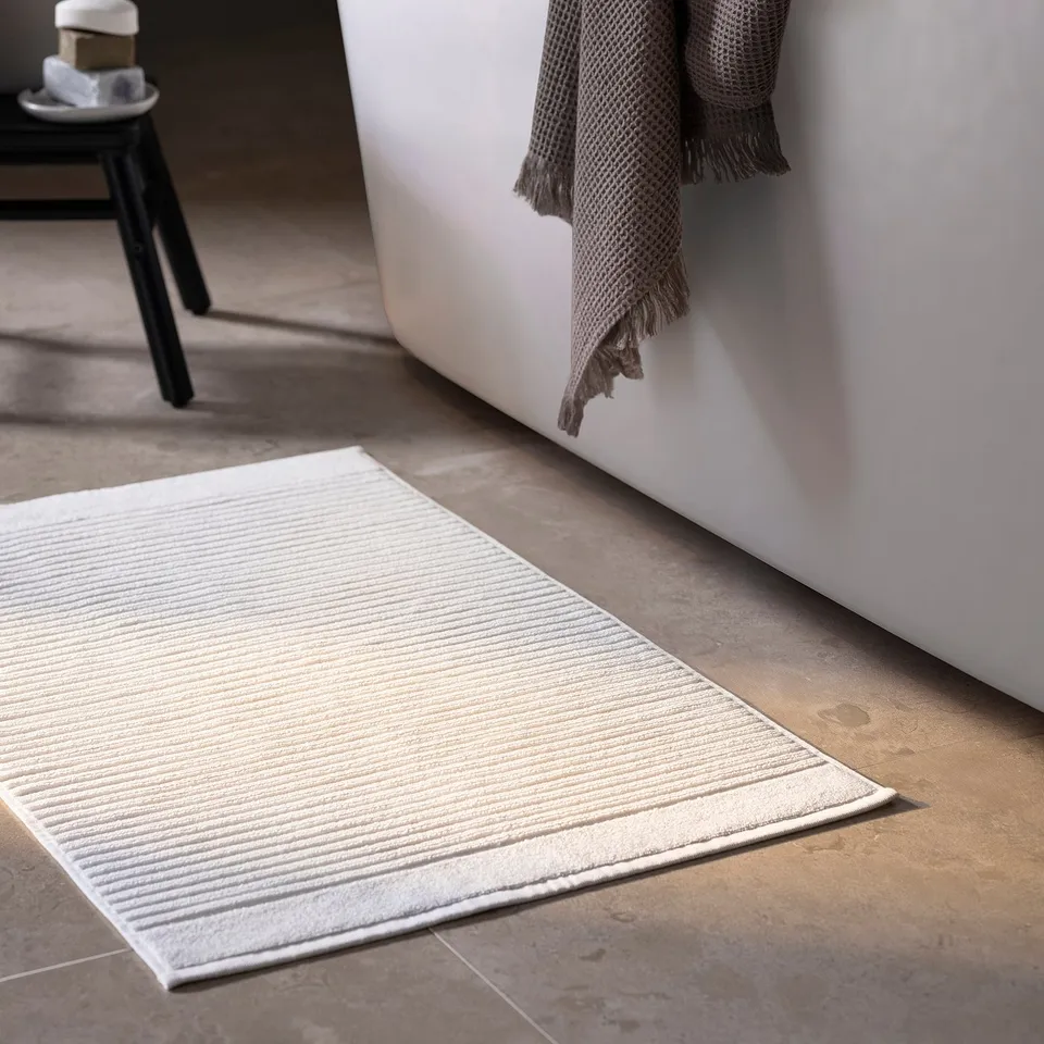 Bathroom with white terry cloth mat, grey towel, black stool.