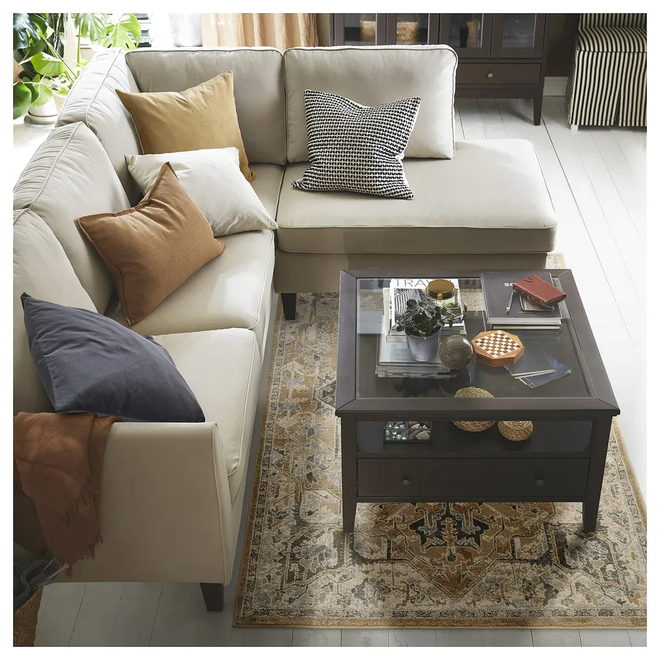 Large cream sectional sofa with colourful pillows, dark coffee table with various items atop, patterned rug beneath, set against light wooden floor.