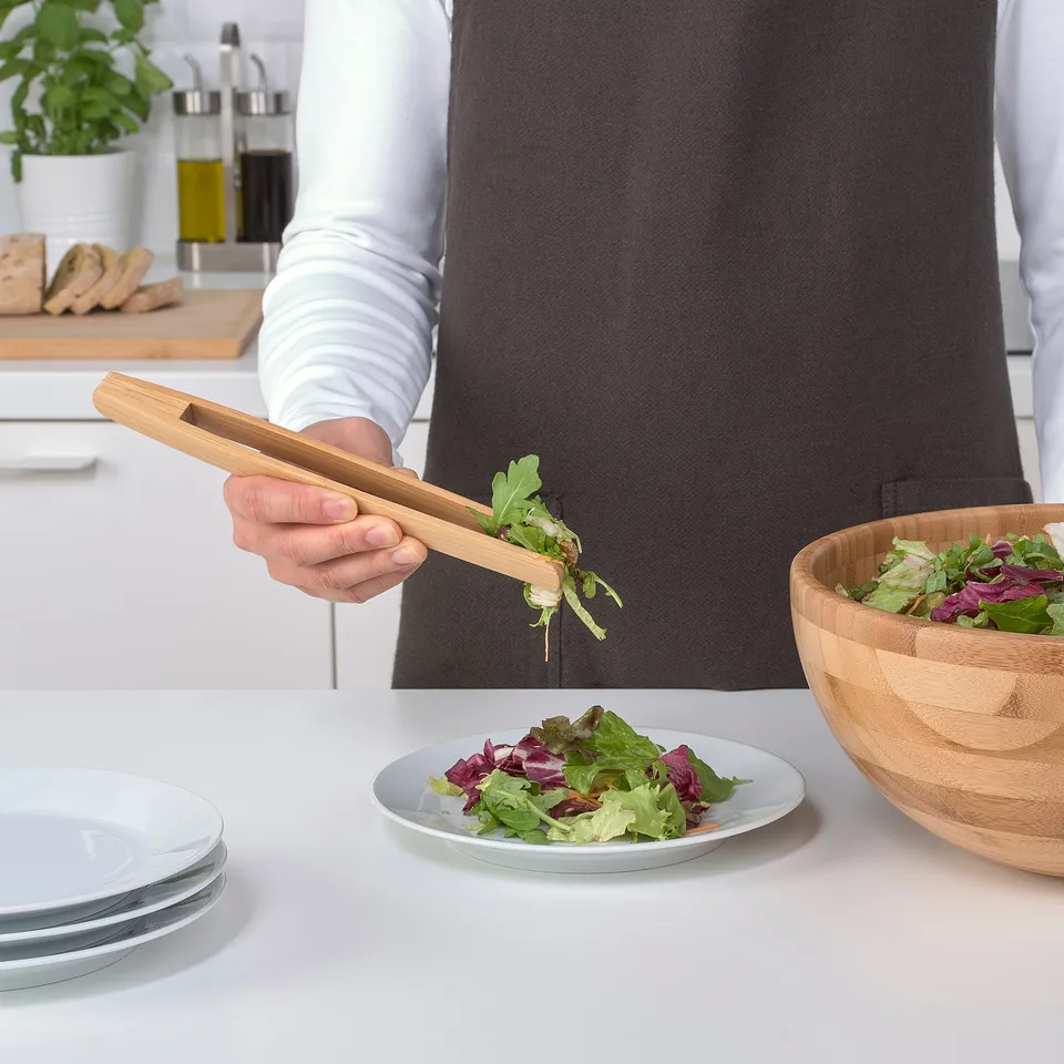 A person is holding a bamboo OSTBIT serving tong, sorting salad greens from a bowl onto a plate. Bamboo tongs, practical for picking and serving food.