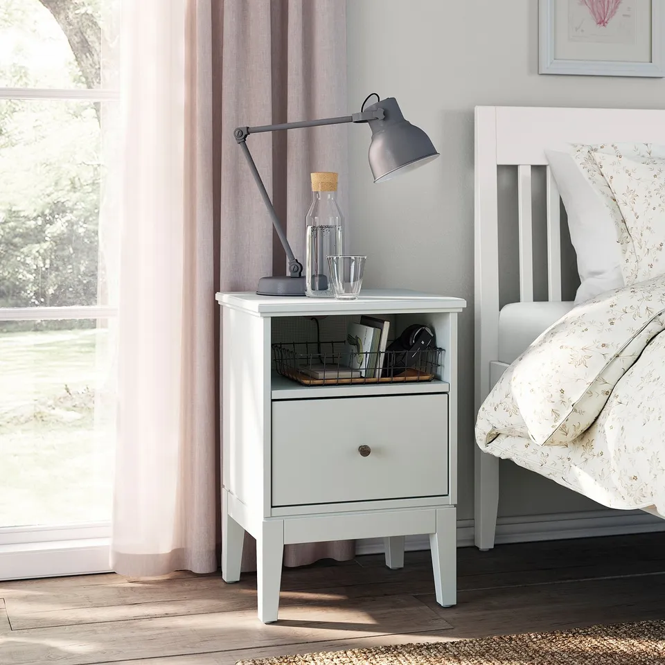 IDANÄS bedside table with lamp, bottle, and basket beside a bed with floral pillows.