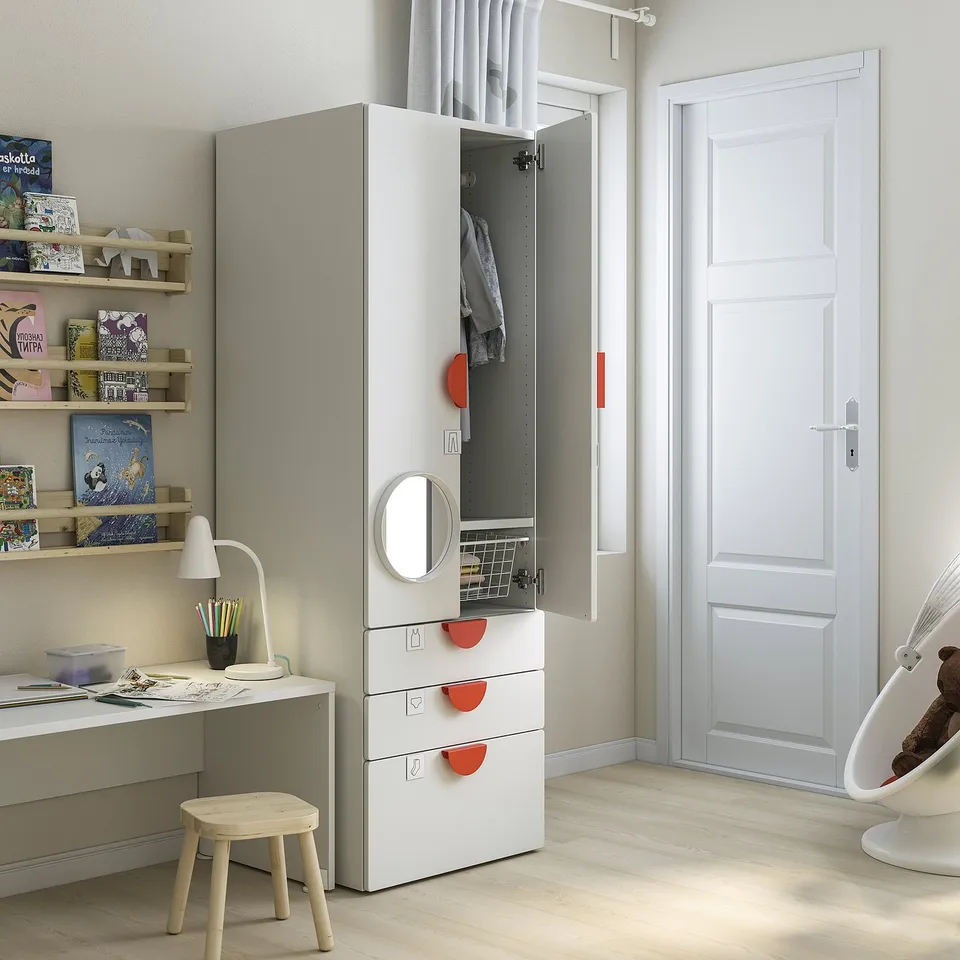 A childs room with a white SMÅSTAD wardrobe, shelves, and desk. The wardrobe has red handles and an open door revealing clothes.