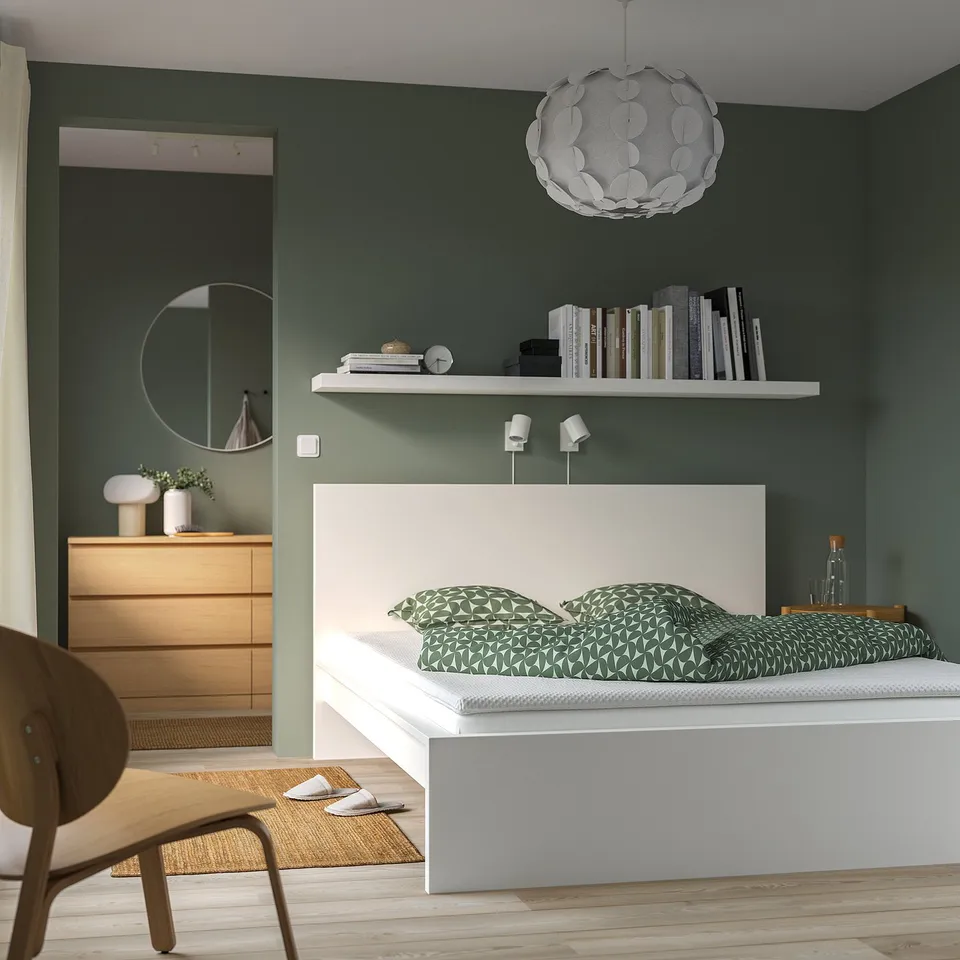 Modern green bedroom with white bed, wooden dresser, and hanging light. Bookshelf above bed.