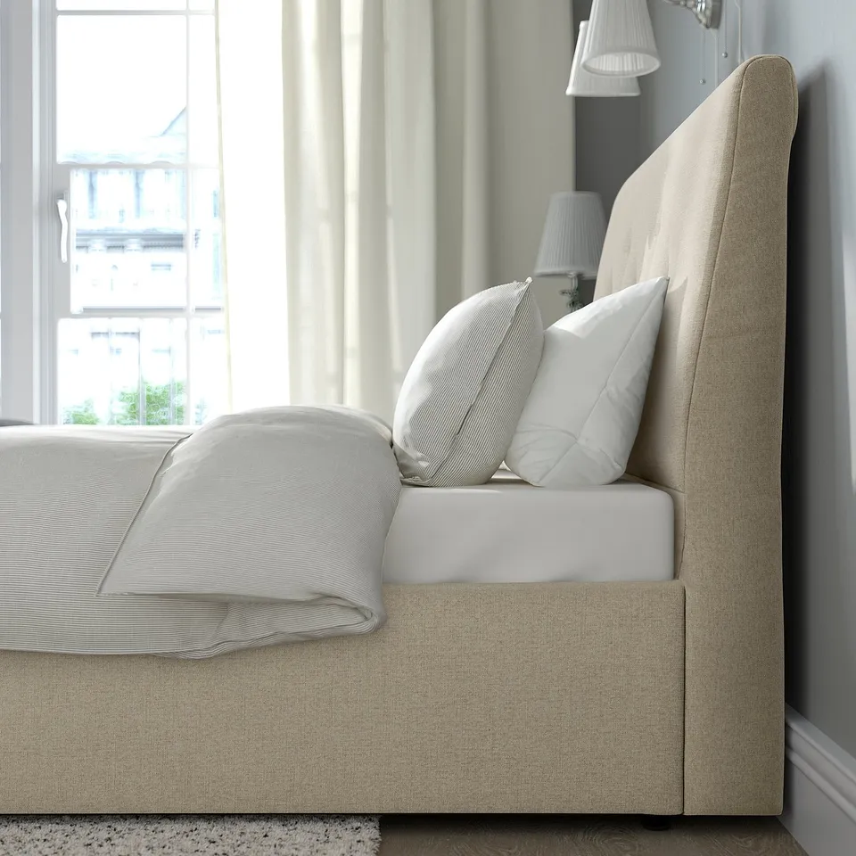Neutral bedroom scene featuring IDANÄS upholstered bed with sloping headboard, soft pillows, and lamps, showcasing built-in storage under the bed.