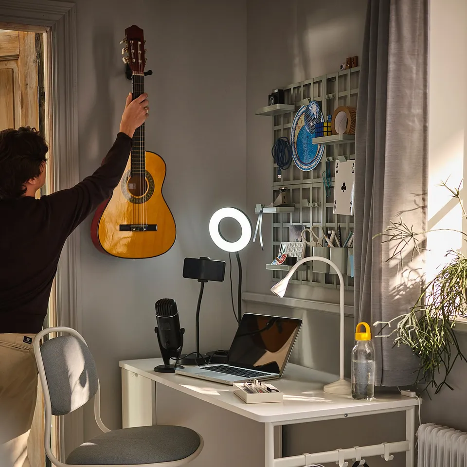 A person tunes a guitar beside a home office setup with a white adjustable chair, desk, and recording equipment.