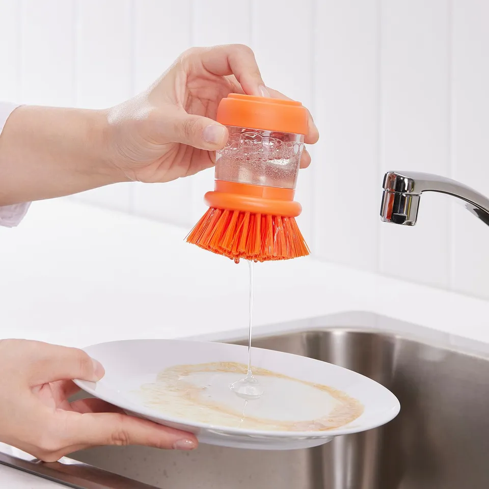 Person squeezing orange brush, releasing soap onto dish. Kitchen sink and tap in background.