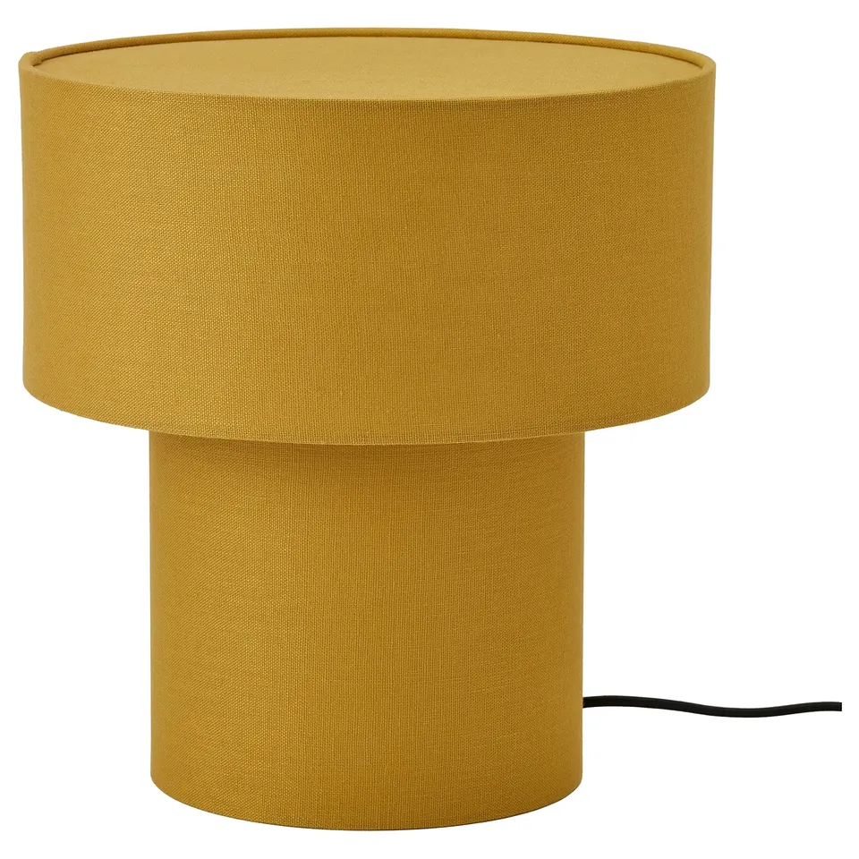 A yellow table lamp named STOCKHOLM 2025, featuring a cylindrical shape and fabric shade, giving a soft warm light.