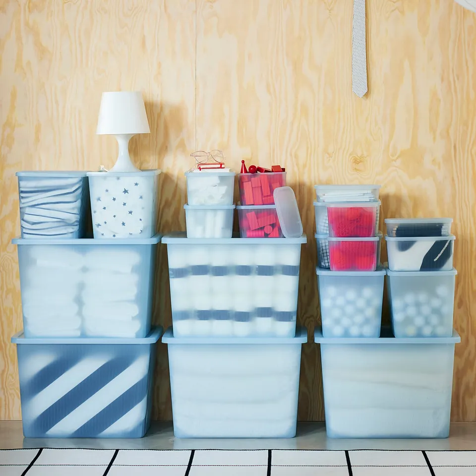 RYKTA boxes in blue, stacked on floor against wooden wall. Various sizes, semi-transparent. Lighting on top.