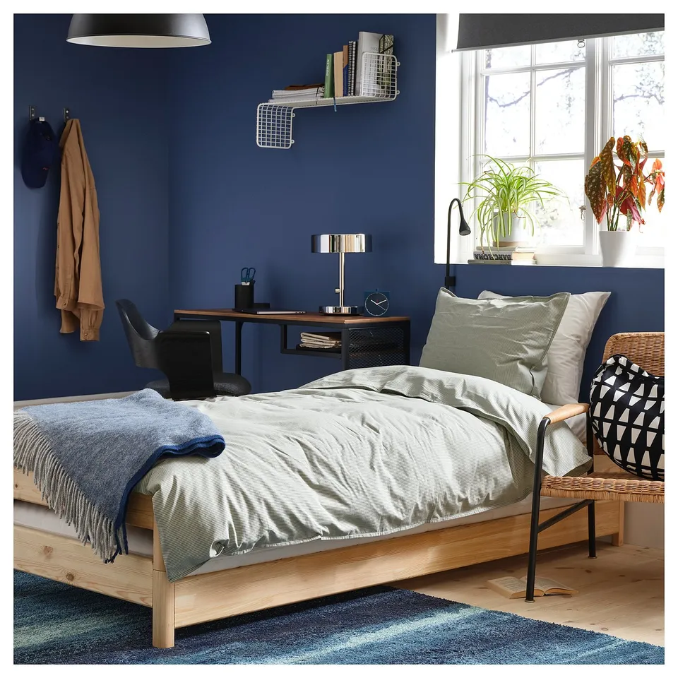 Modern blue bedroom with wooden bed, green pillows, workspace, and wicker chair.
