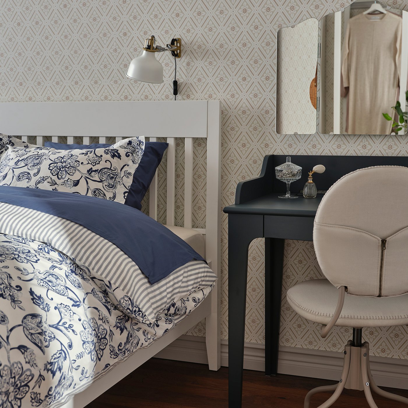 Bedroom with white IDANÄS bed, blue floral print bedding, wall-mounted lamp, dark desk, cream chair, and decorative wallpaper.