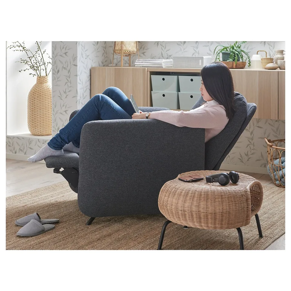 Person in grey recliner with footrest, beside stool with book & headphones, vase with branches, floral wallpaper.