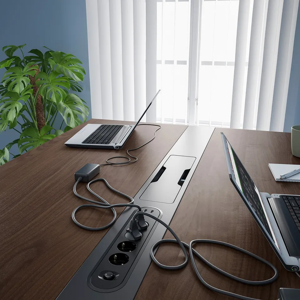 Wooden conference table with laptops, cables, and power module for charging, set against a window.